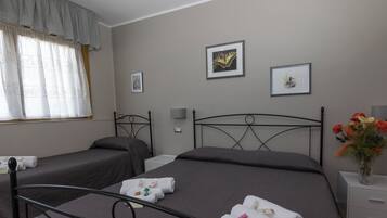 A hotel room with two single beds, a large bed, a window with curtains, a bedside table with a vase of flowers, and wall art.