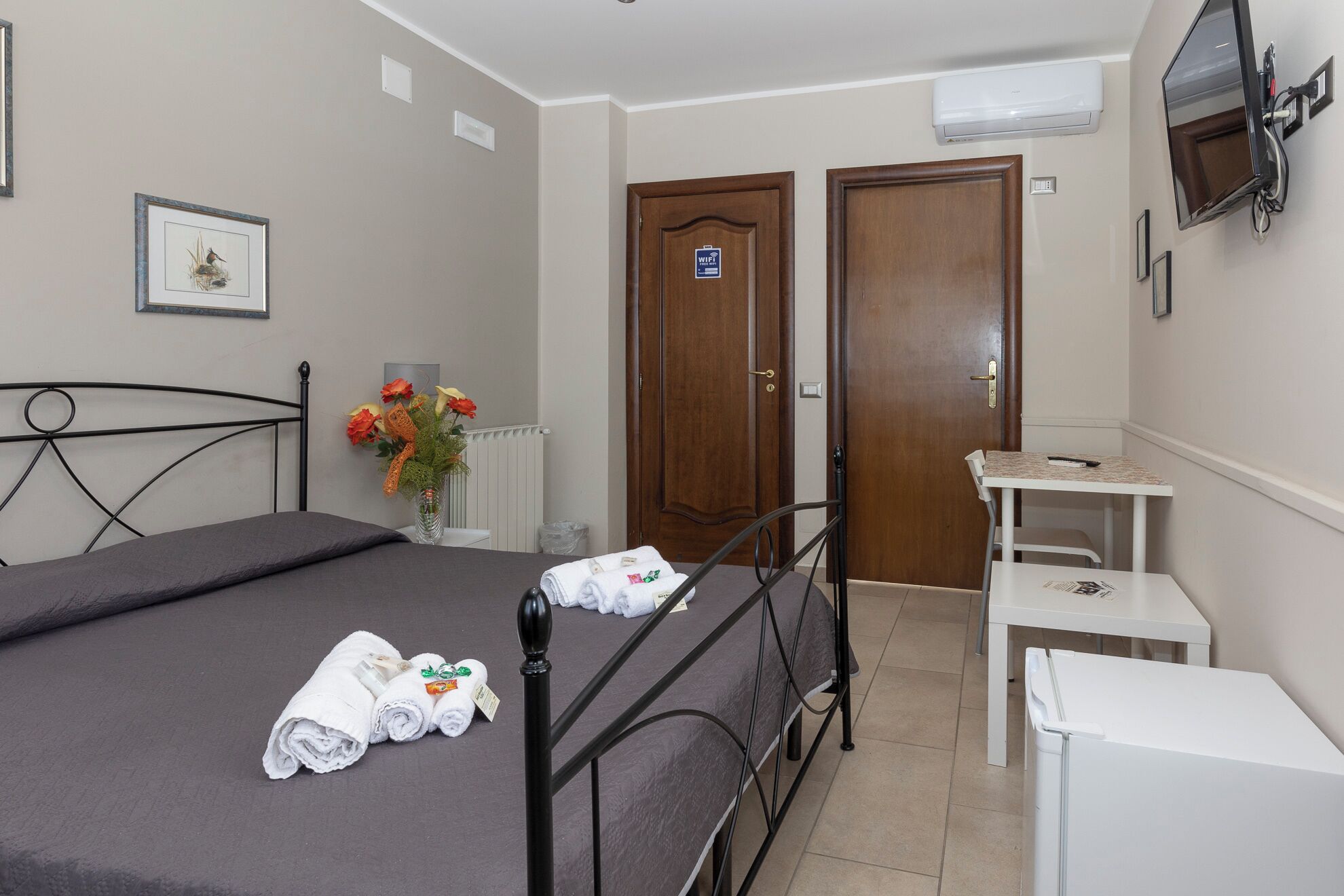 Habitación de hotel con cama, una mesita, una silla, un radiador y televisor montado en la pared.