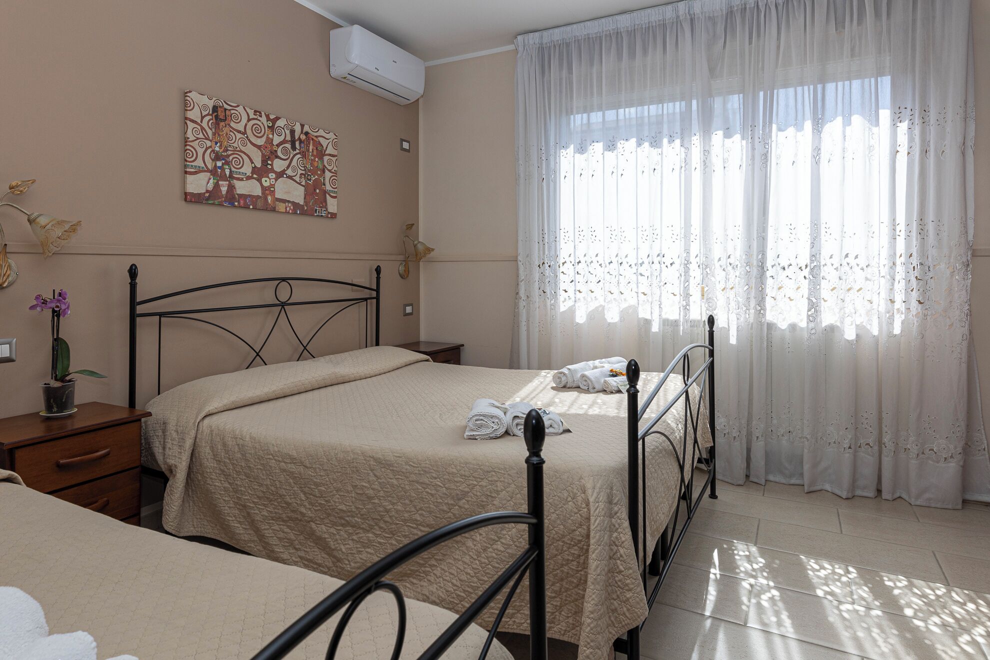 Habitación de hotel con dos camas, un ventanal con cortinas translúcidas, un aire acondicionado de pared y un cuadro decorativo.