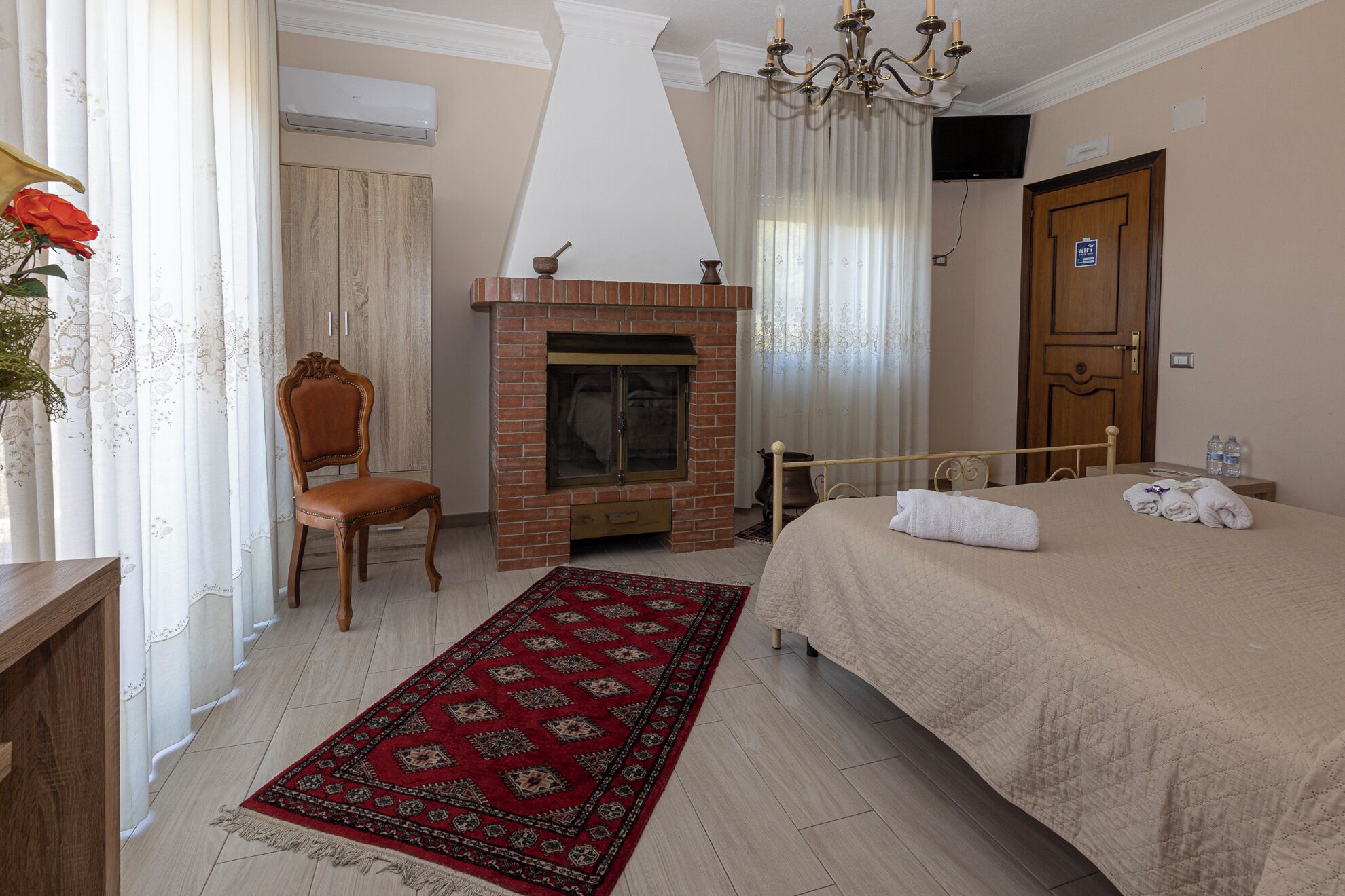 Una habitación de hotel con cama, una silla de madera, chimenea, alfombra roja estampada y un candelabro.