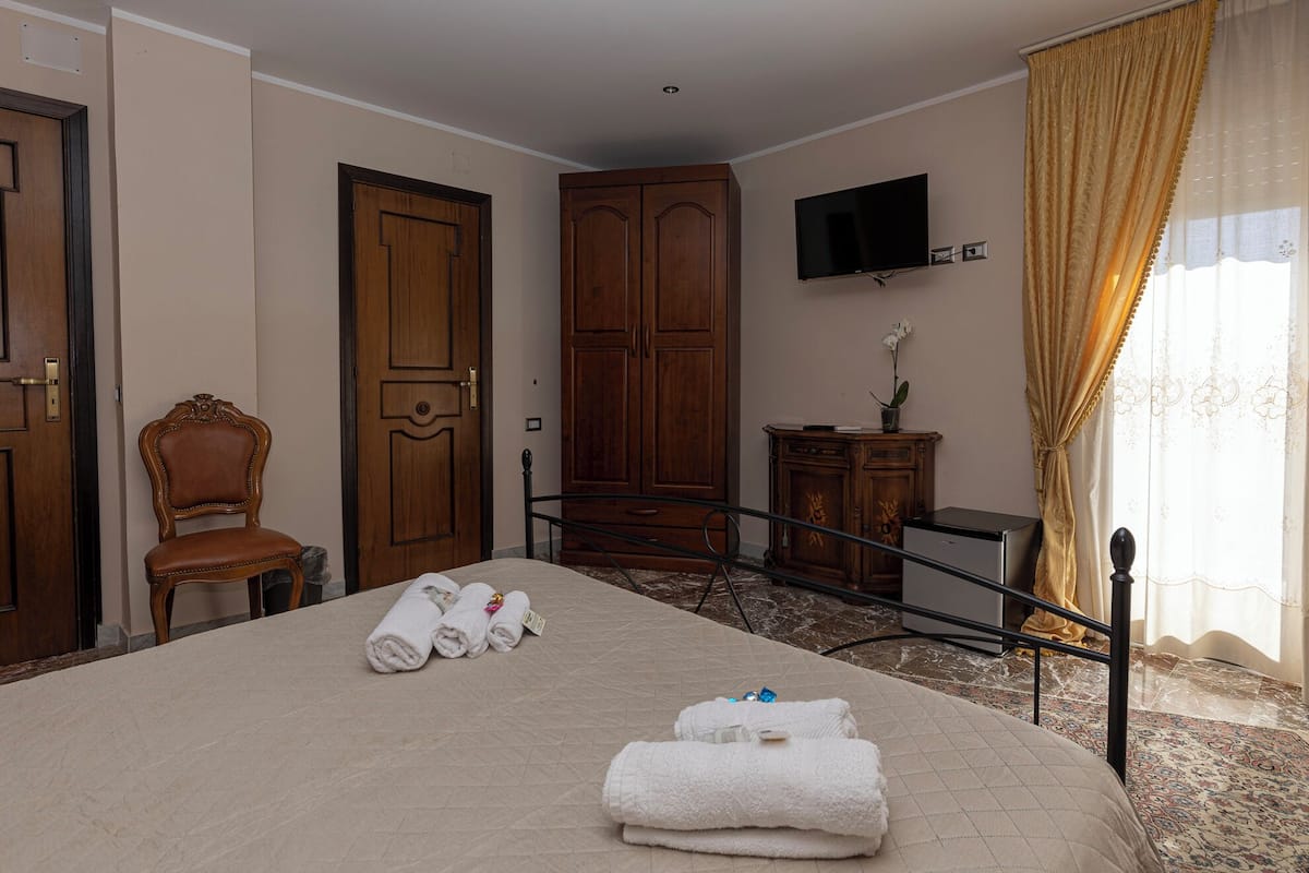 A hotel room with a bed, a chair, a wardrobe, a television, and a desk with a plant.