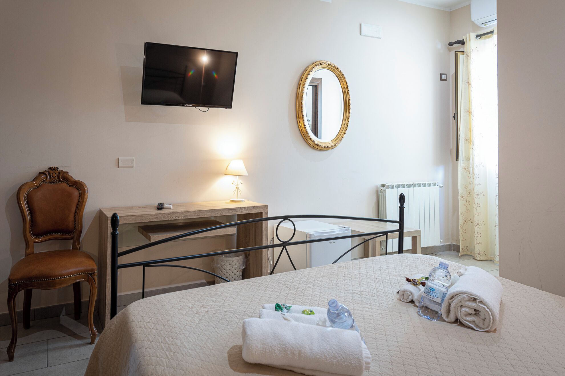 Quarto de hotel com cama, poltrona, escrivaninha com abajur, televisão, espelho e radiador.