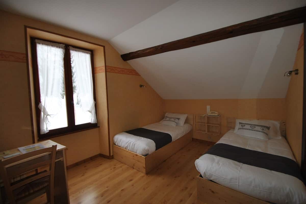 Une petite chambre mansardée, chaleureuse et accueillante, avec deux lits en bois, un bureau et une fenêtre avec des rideaux.