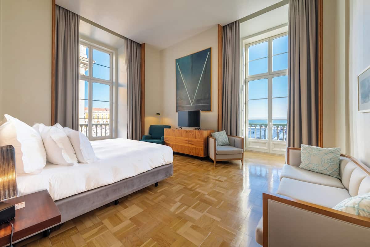 Habitación de hotel con una cama grande, un sofá, un escritorio y un televisor. Cuenta con ventanas amplias con cortinas y un cuadro en la pared.