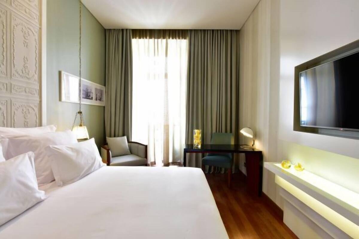 Habitación de hotel con una cama grande, un escritorio con una silla, un televisor y un ventanal con cortinas.