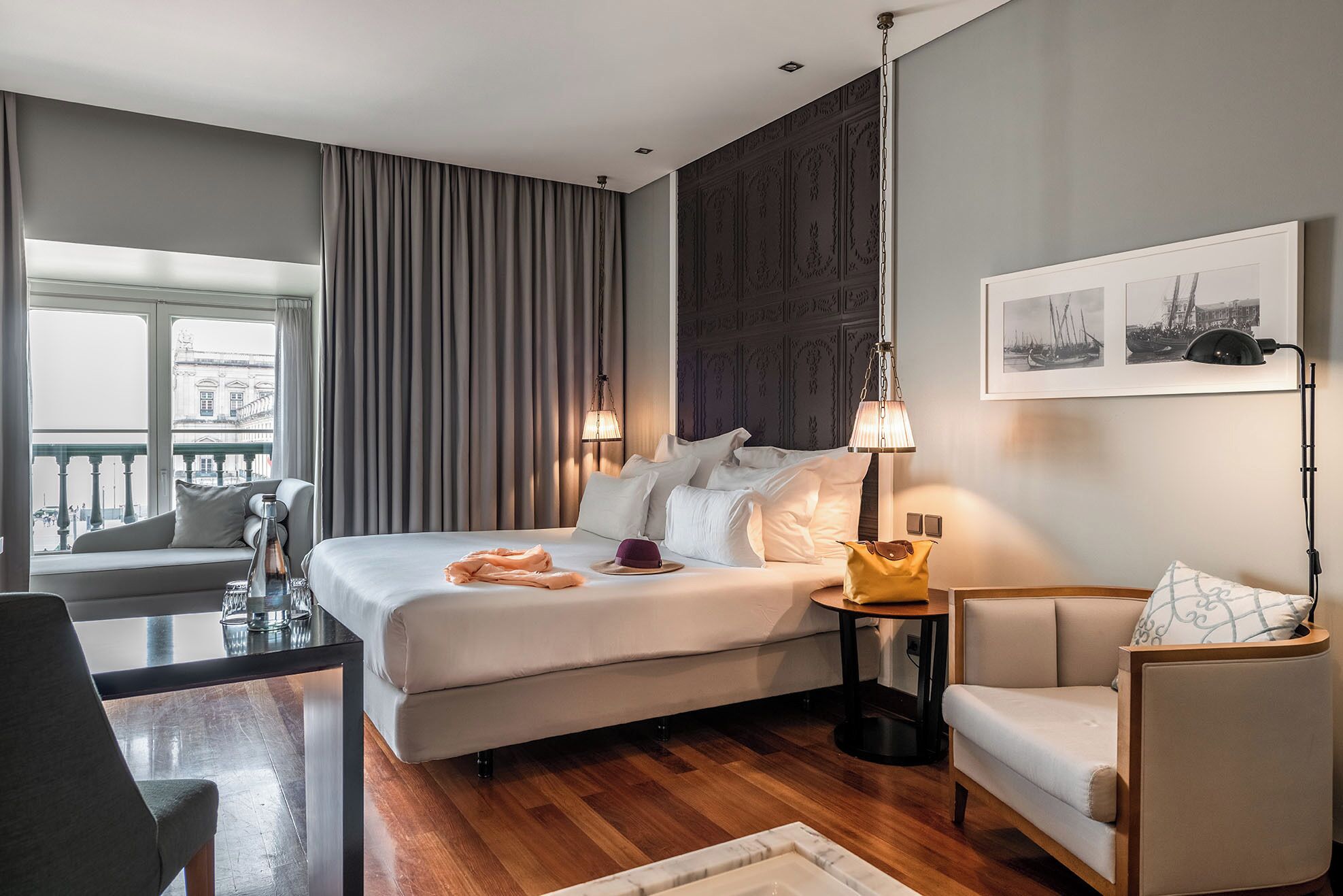 Una habitación de hotel moderna con una cama grande, un área de descanso con sofá y sillón, una mesita y vistas a la ciudad a través de la ventana.