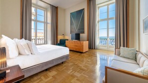 Una habitación de hotel con cama, sofá, televisión, cómoda de madera y ventanas amplias con cortinas.