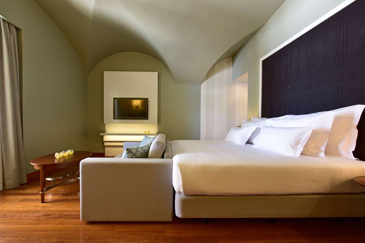 Una habitación de hotel moderna con una cama grande, un sofá, una mesita y un televisor montado en la pared.