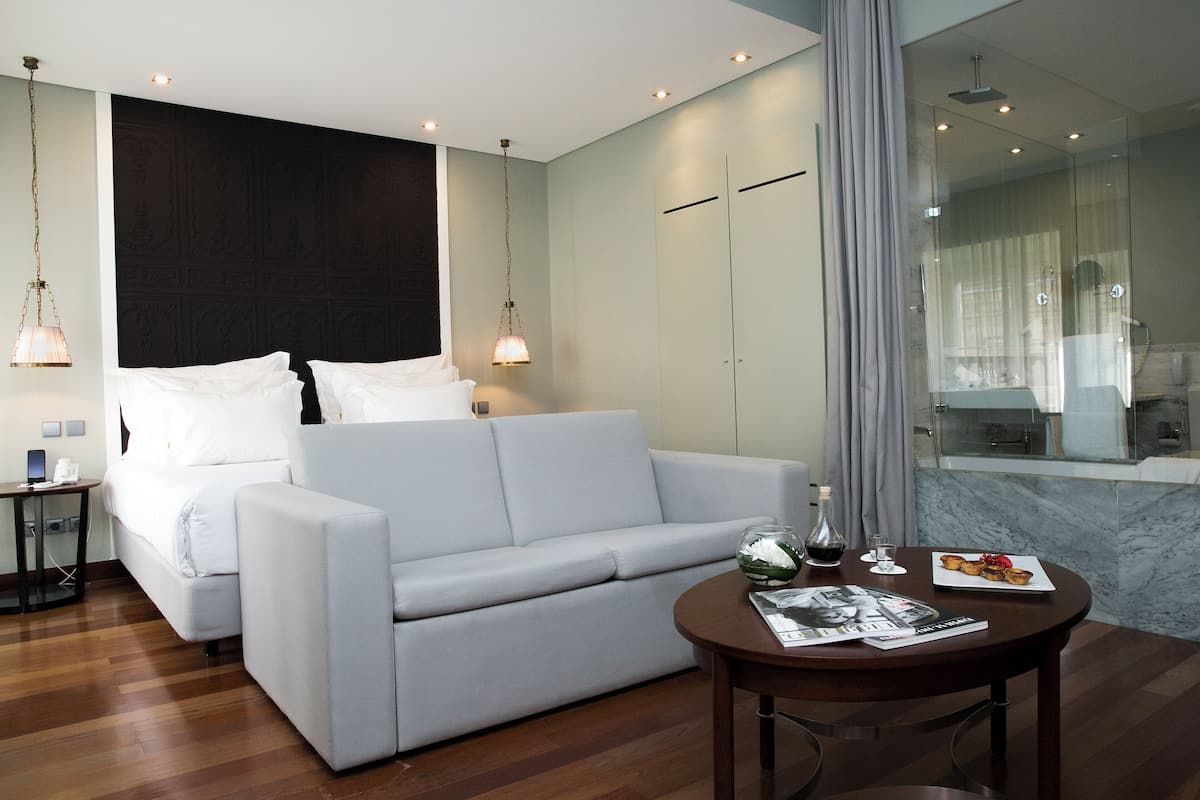 Habitación de hotel moderna con un sofá blanco, una mesita redonda con una revista y una copa de vino, y un ventanal con vistas al baño.