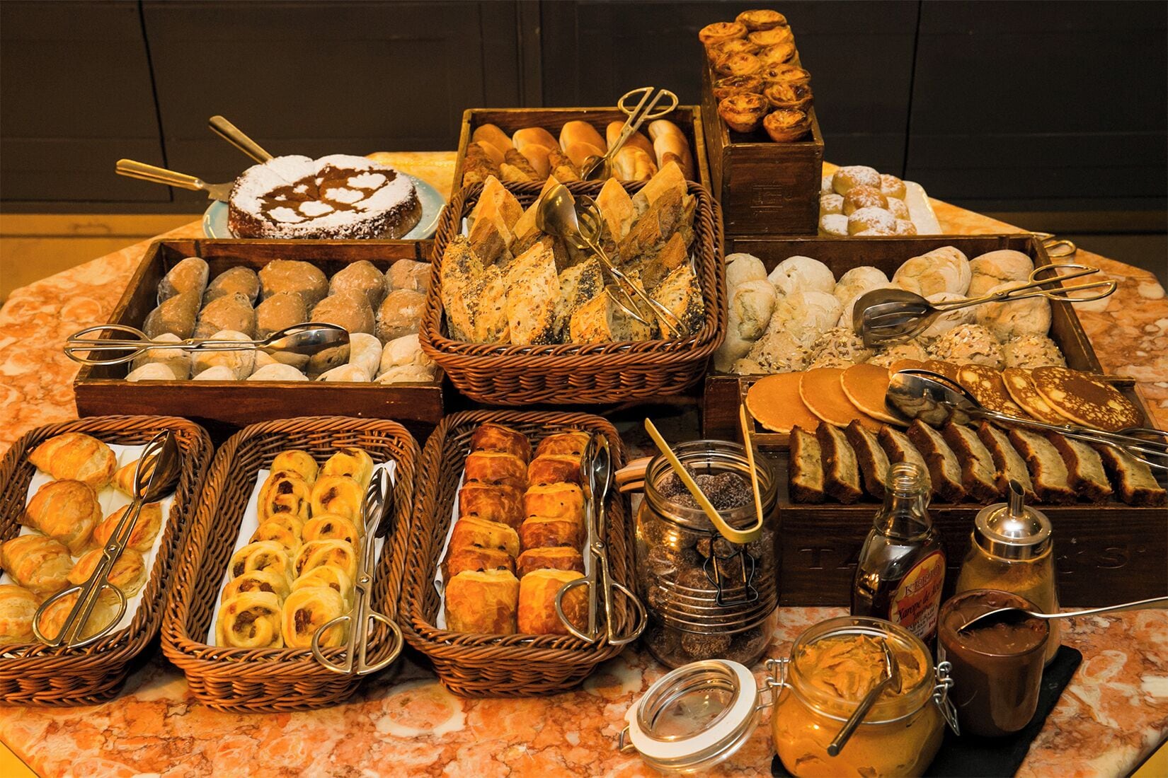 Une variété de produits de boulangerie et de pâtisserie présentés dans des paniers et sur des plateaux, accompagnés d’ustensiles pour servir.