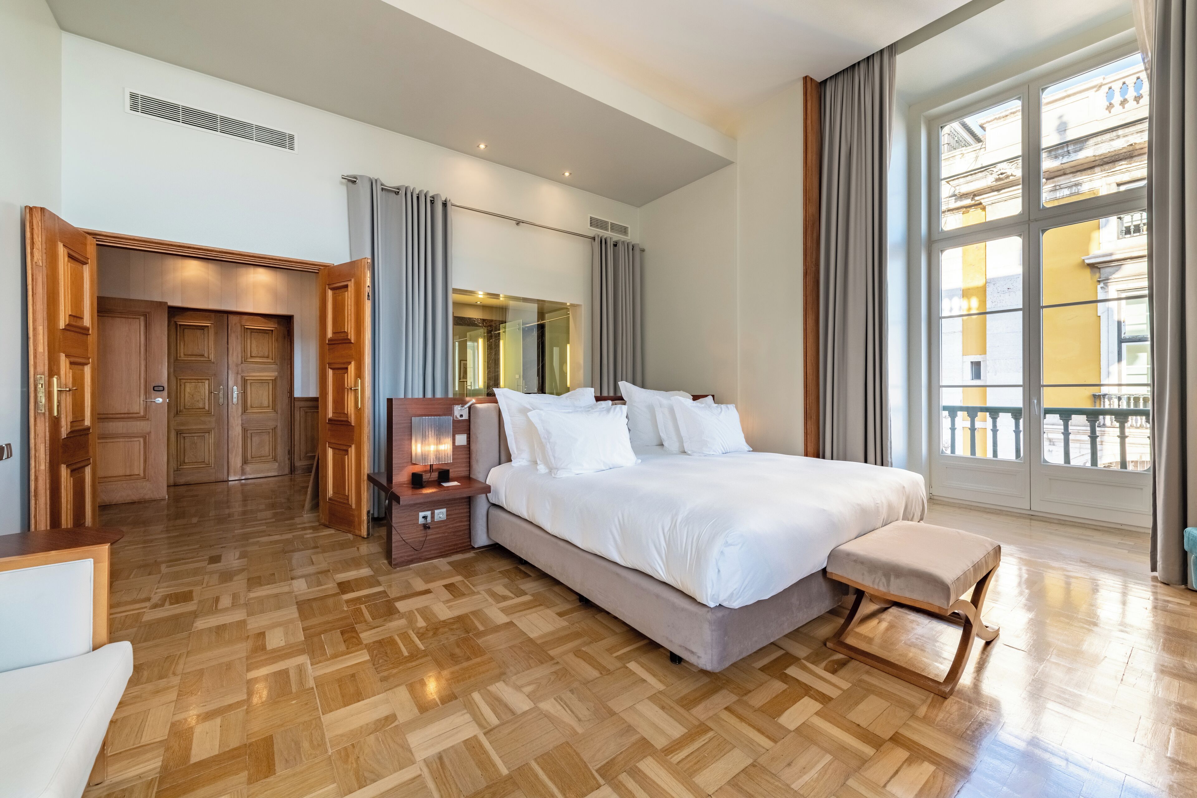 A spacious bedroom with a large bed, a wooden floor, a nightstand with a lamp, and a view of the city through large windows.
