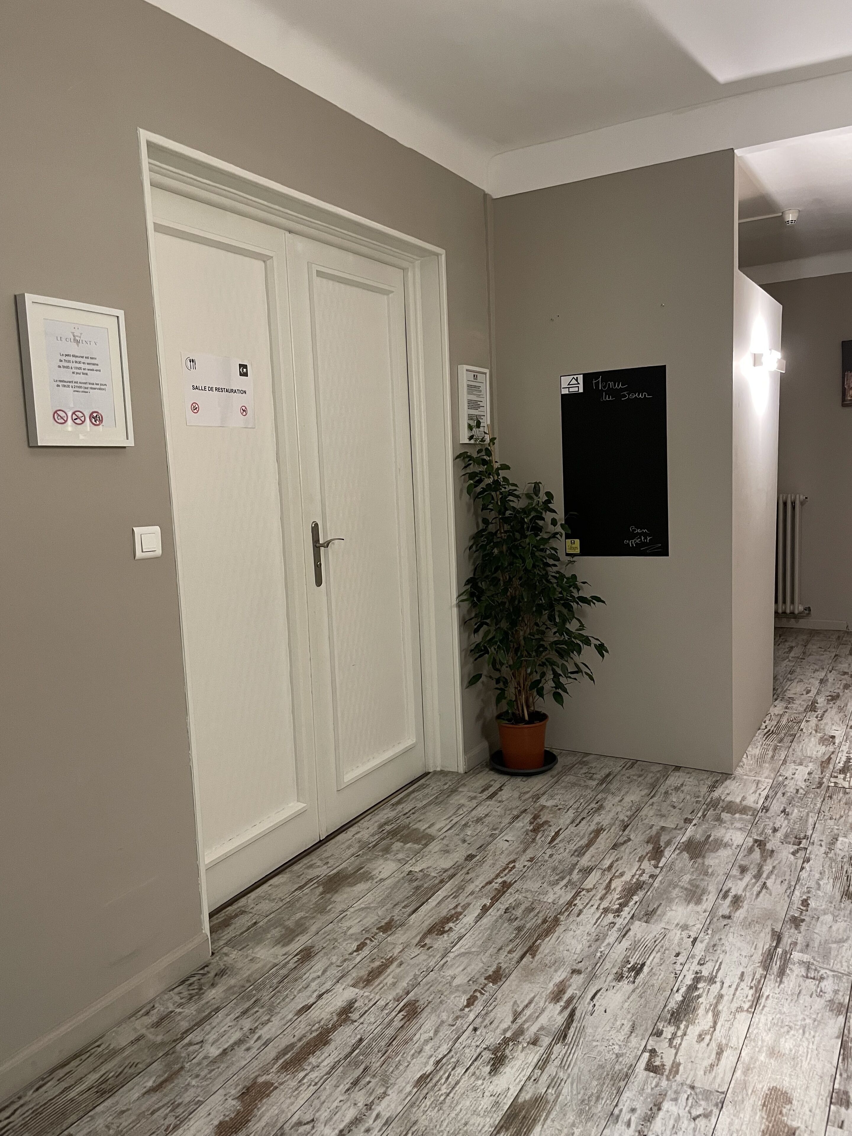 A hallway with a white door, a potted plant, and signs indicating a restaurant and a menu board.