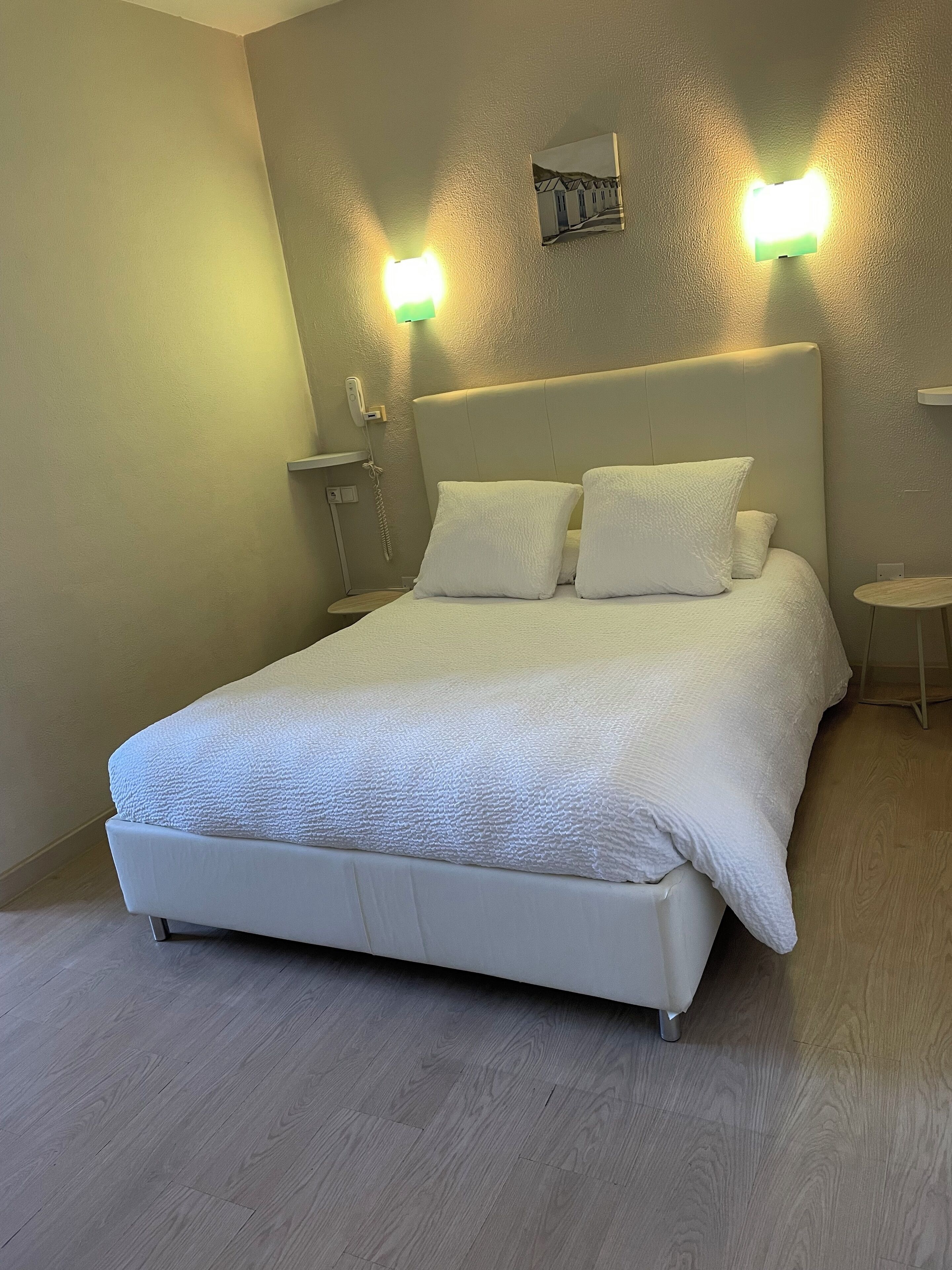 A neatly made bed with a white comforter and pillows, a bedside table with a lamp, and a wall-mounted phone.