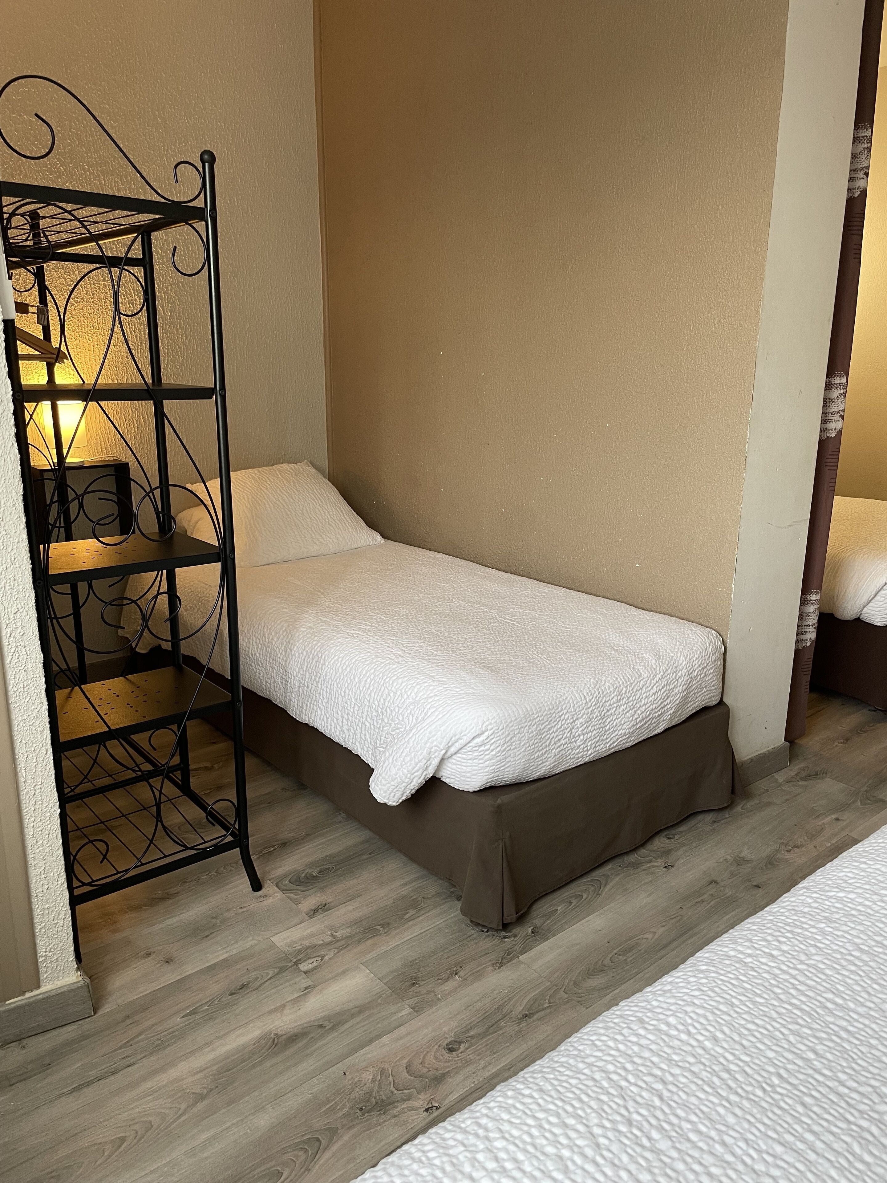 A small, single-bed room with a metal shelf unit against the wall, a bedside table, and a lamp.
