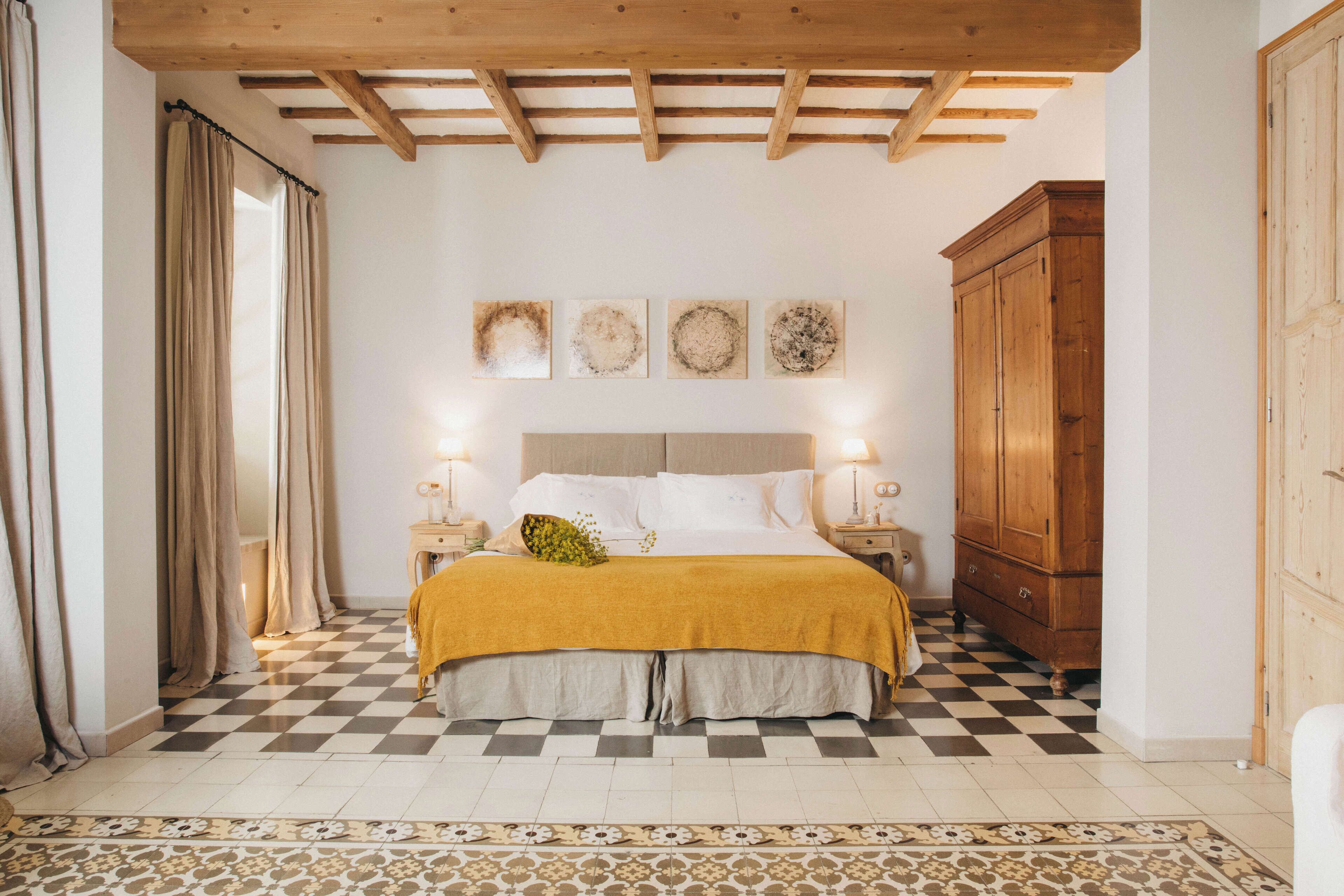 Un dormitorio con piso a cuadros, cama con manta color mostaza, cabecera de madera, lámparas de noche y un armario de madera.