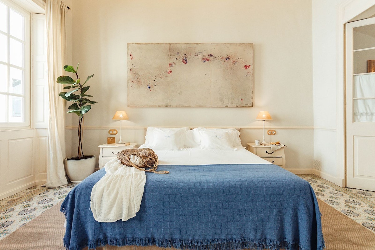 A bedroom with a large bed, two bedside tables, a potted plant, and a painting on the wall.