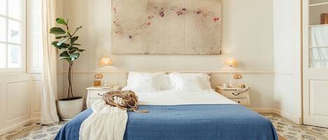 A bedroom with a large bed, two bedside tables, a potted plant, and a painting on the wall.