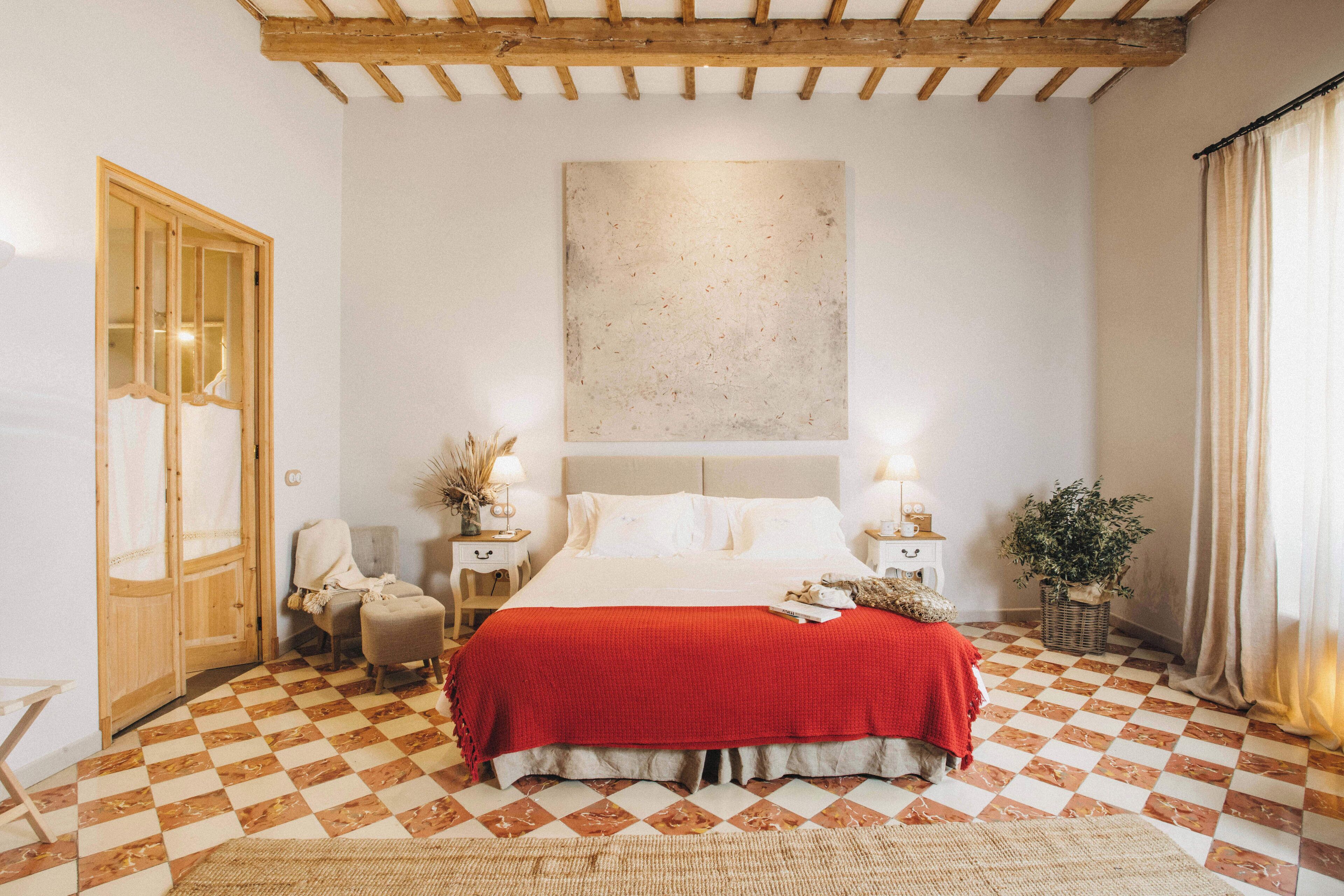 A bedroom with a checkered floor, a large bed with a red blanket, two bedside tables with lamps, a wooden door, and a large abstract painting on the wall.