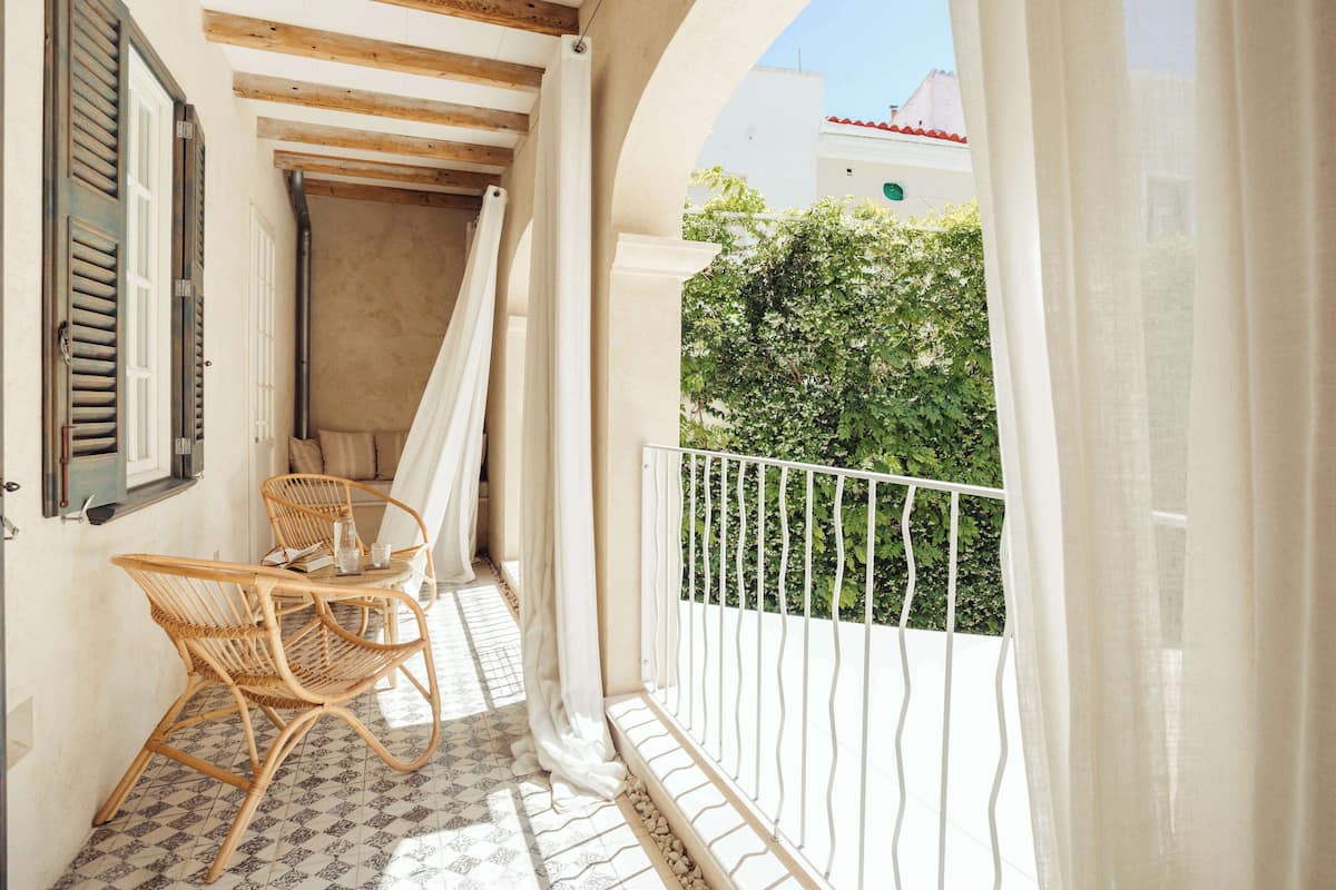 A balcony with wicker chairs, a white balustrade, and a view of greenery and a building with a red roof.
