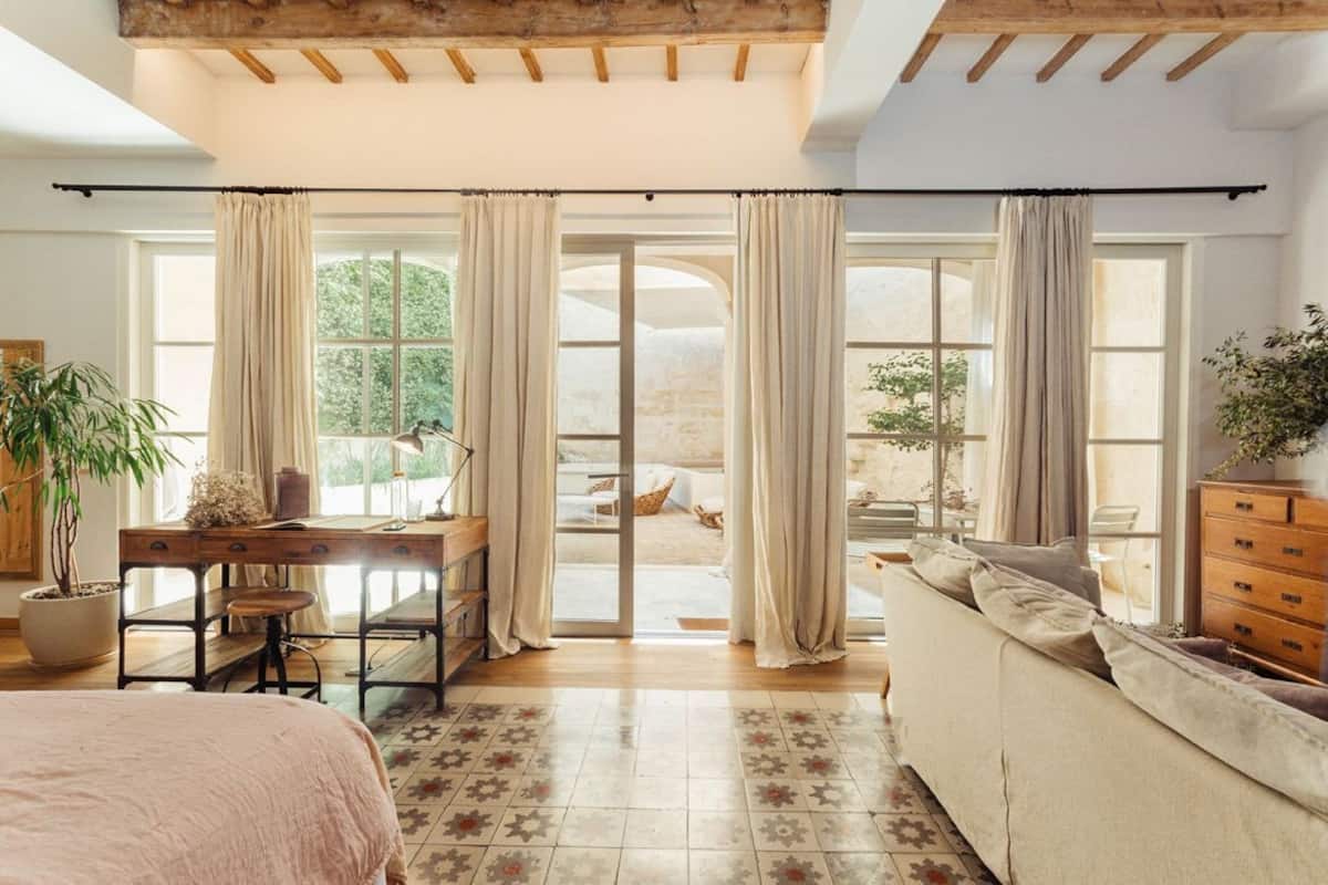 A bedroom with a bed, a desk, a sofa, and a view of the outdoors through large windows.