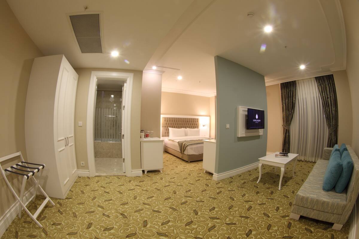 A hotel room with a patterned carpet, a bed with a headboard, a TV mounted on the wall, a white sofa with blue cushions, and a small table.