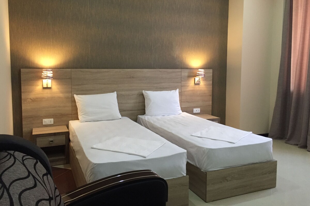 A hotel room with two single beds, wooden headboards, bedside lamps, and a small nightstand.