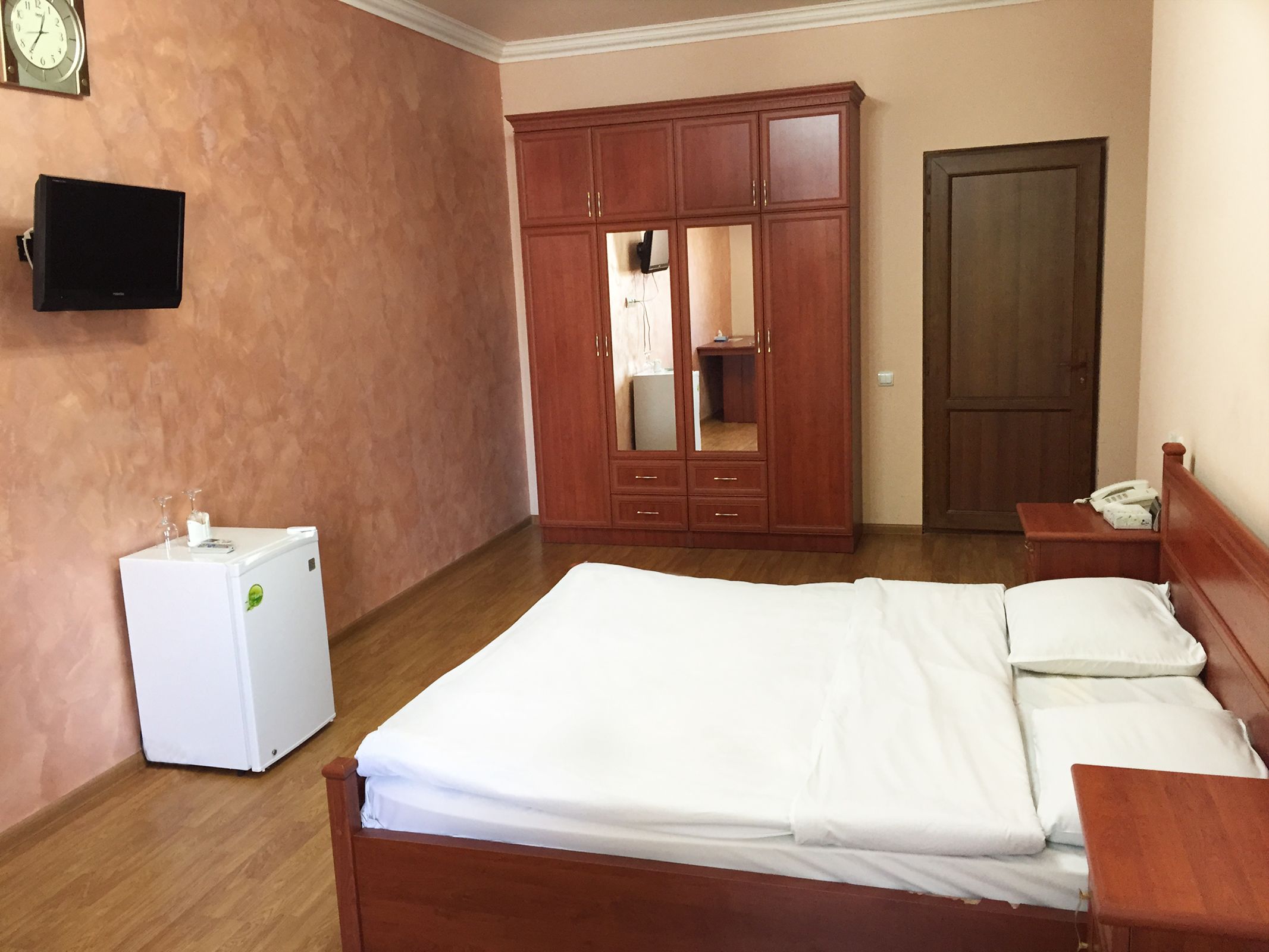Un dormitorio con cama, armario, refrigerador pequeño, mesita de noche con teléfono y televisor montado en la pared.