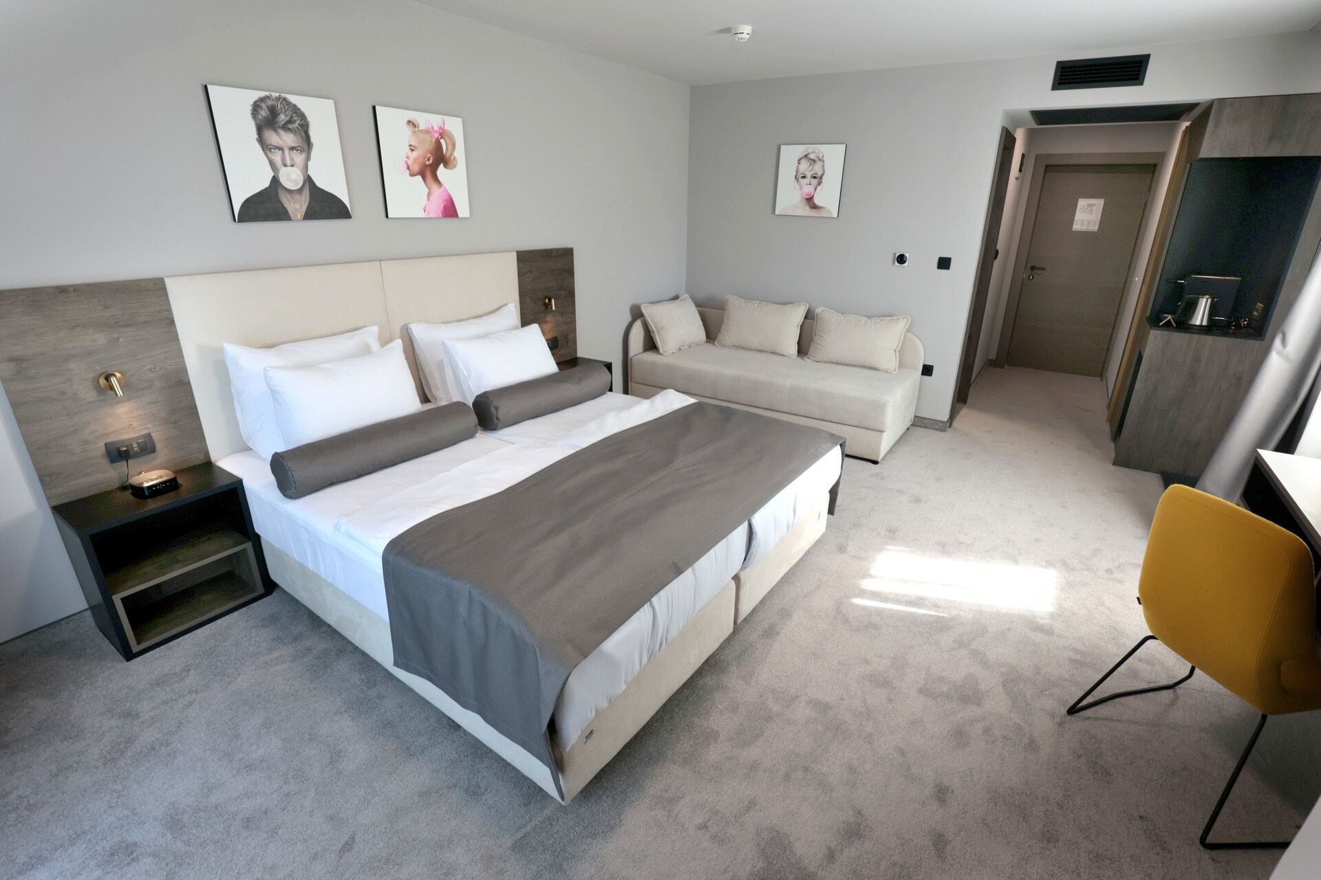 A modern hotel room with a large bed, a sofa, a desk, and a chair. There are three framed pictures on the wall.