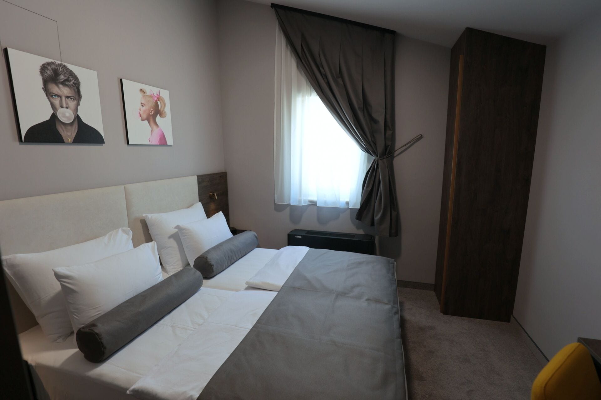 Habitació d'hotel amb un llit, una taula de nit, una finestra amb cortines i dos quadres al mur.