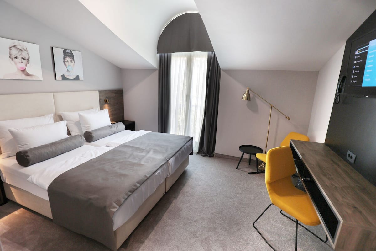 A modern hotel room with a large bed, a desk, and a chair. There is a TV on the wall and a window with curtains.