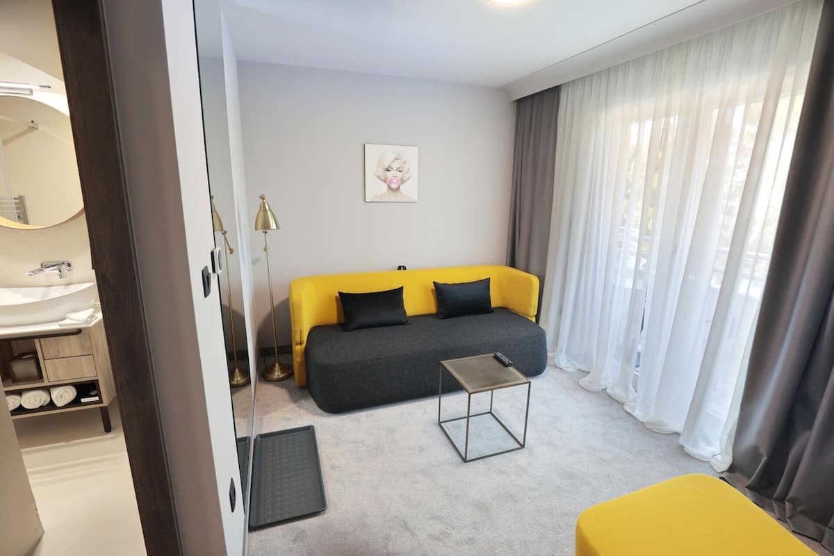 A modern hotel room with a sofa, a small table, a mirror, and a window with curtains.