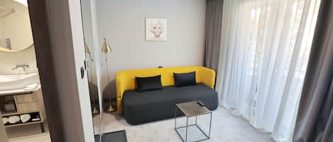 A modern hotel room with a sofa, a small table, a mirror, and a window with curtains.