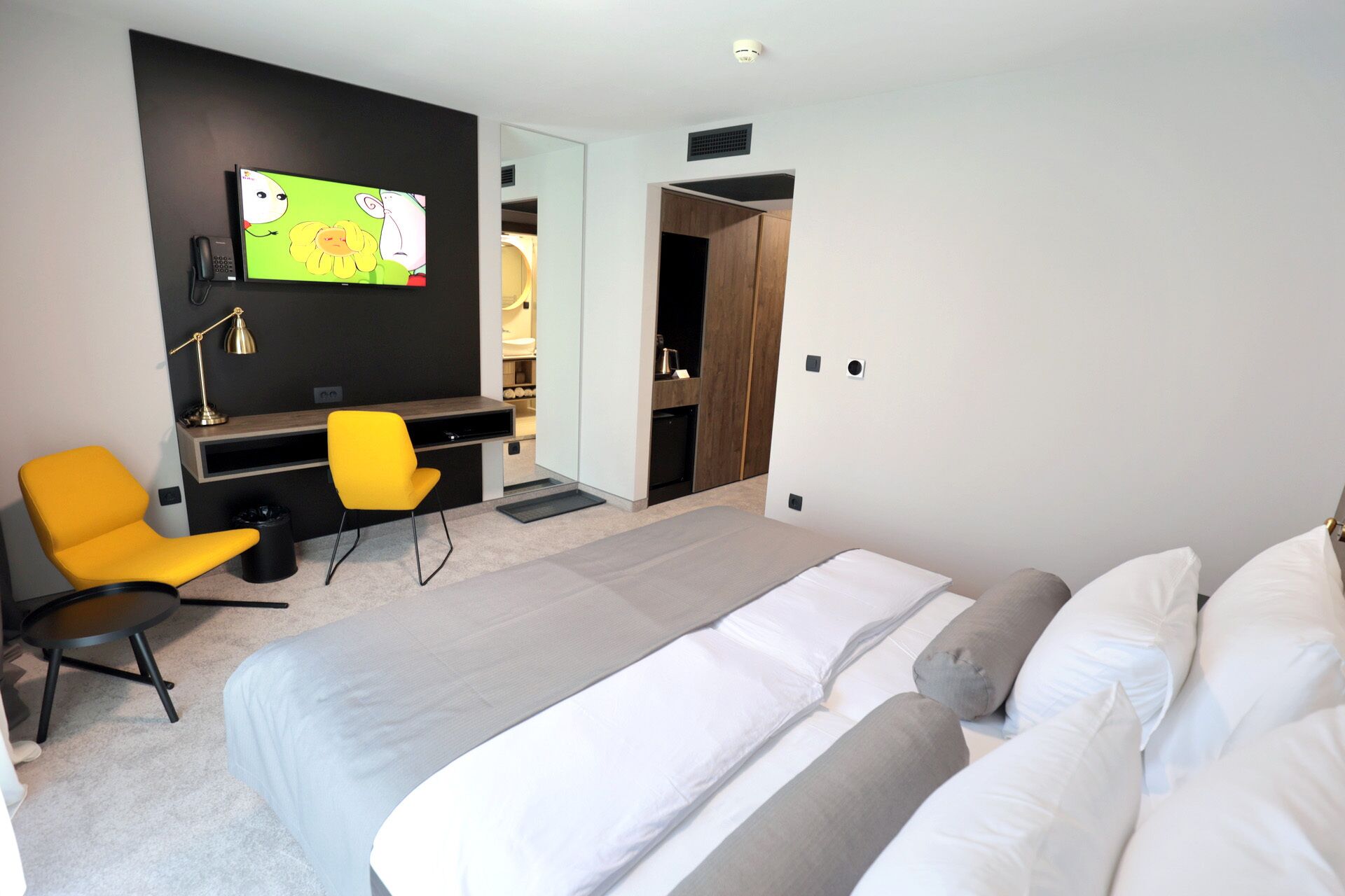 A modern hotel room with a large bed, a desk with a chair, a television, and a wardrobe.
