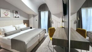 A modern hotel room with a large bed, a desk, and two chairs. There are curtains, a flat-screen TV mounted on the wall, and a ceiling light. - Hotel Milenij (Makarska)