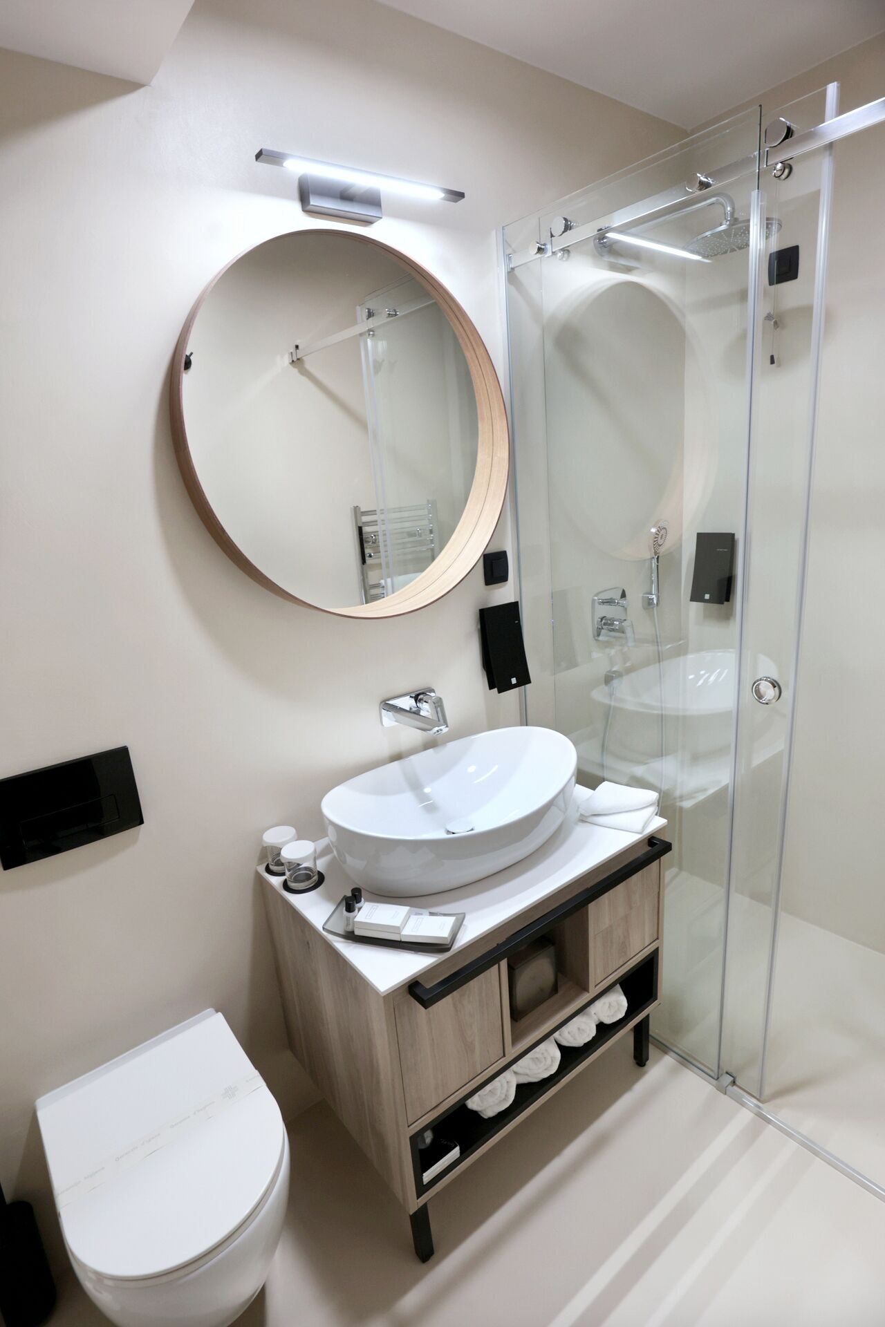 A modern bathroom with a round mirror, a glass-enclosed shower, a white sink, and a toilet.