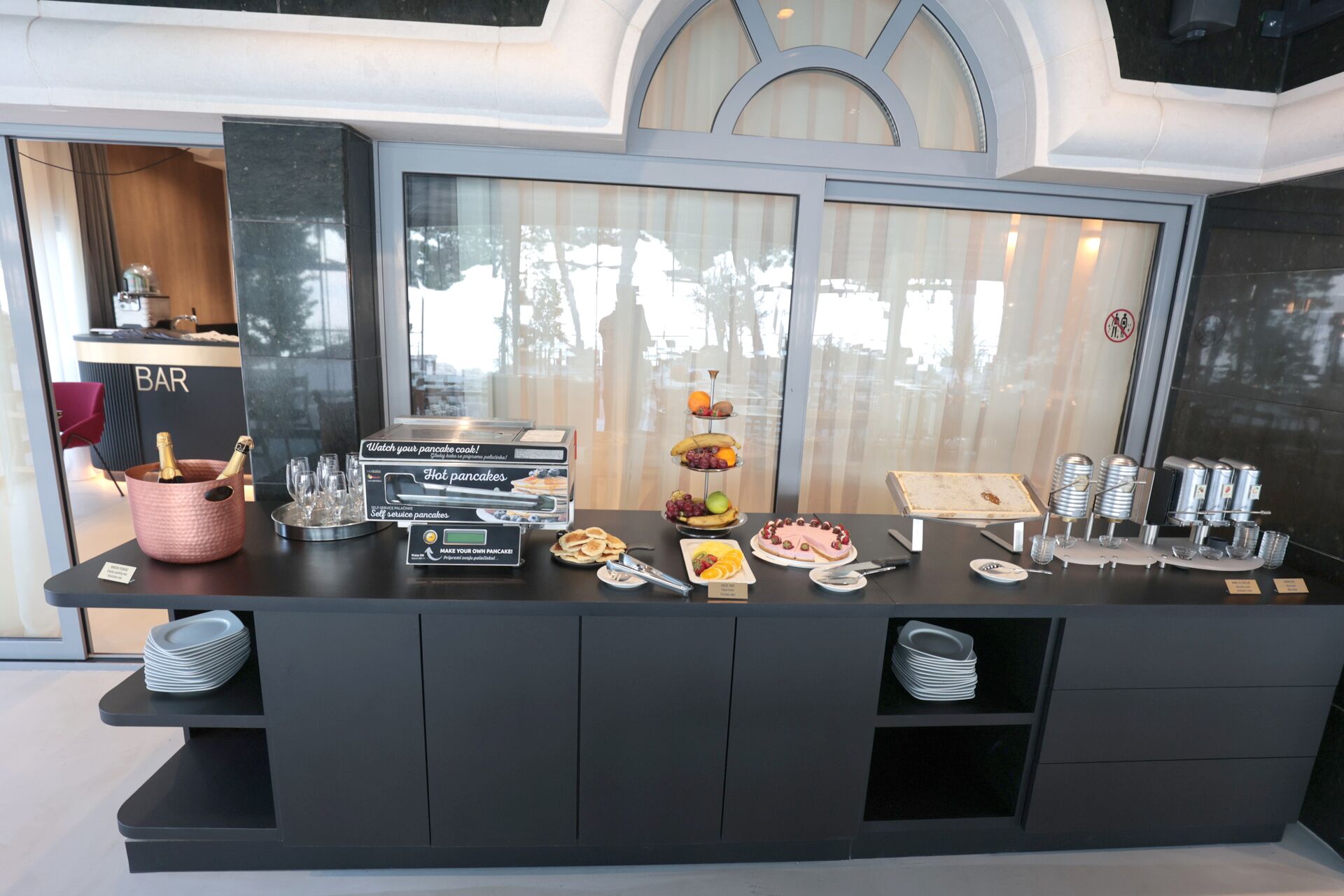 A breakfast bar with a self-service pancake station, a bar area, and a display of breakfast items including pancakes, fruits, and pastries.