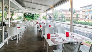 Un amplio espacio para comer al aire libre con mesas y sillas blancas, grandes ventanales que ofrecen vistas a edificios y áreas verdes.