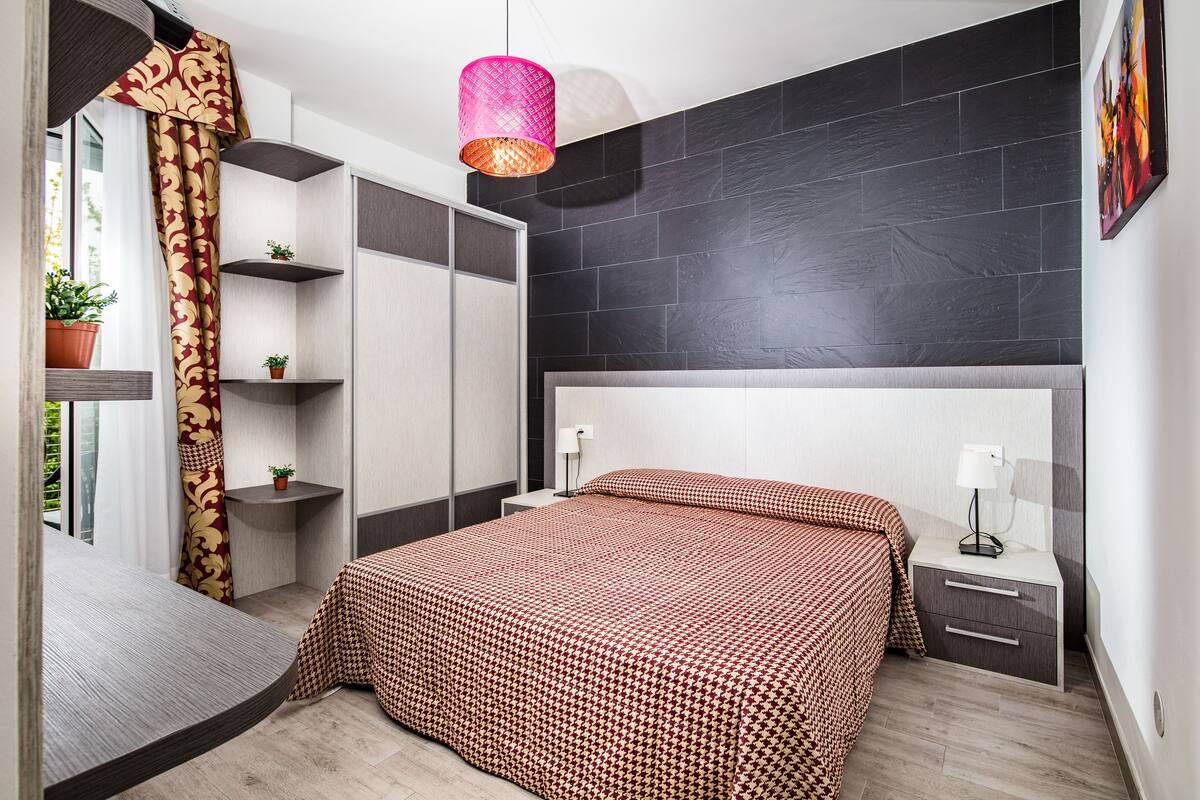 A modern bedroom with a large bed, a bedside table, a nightstand, a hanging light fixture, and a window with curtains.