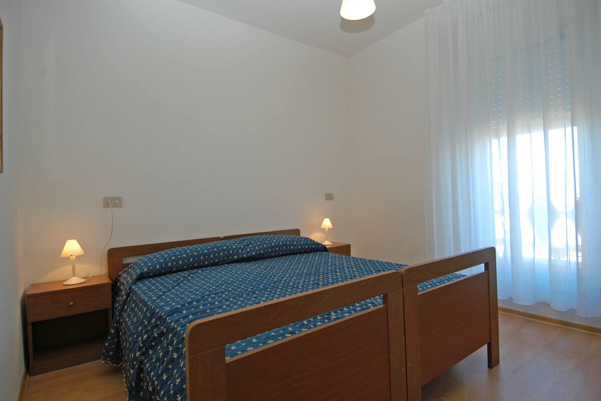 A bedroom with a wooden bed, two bedside tables with lamps, and a window with sheer curtains.