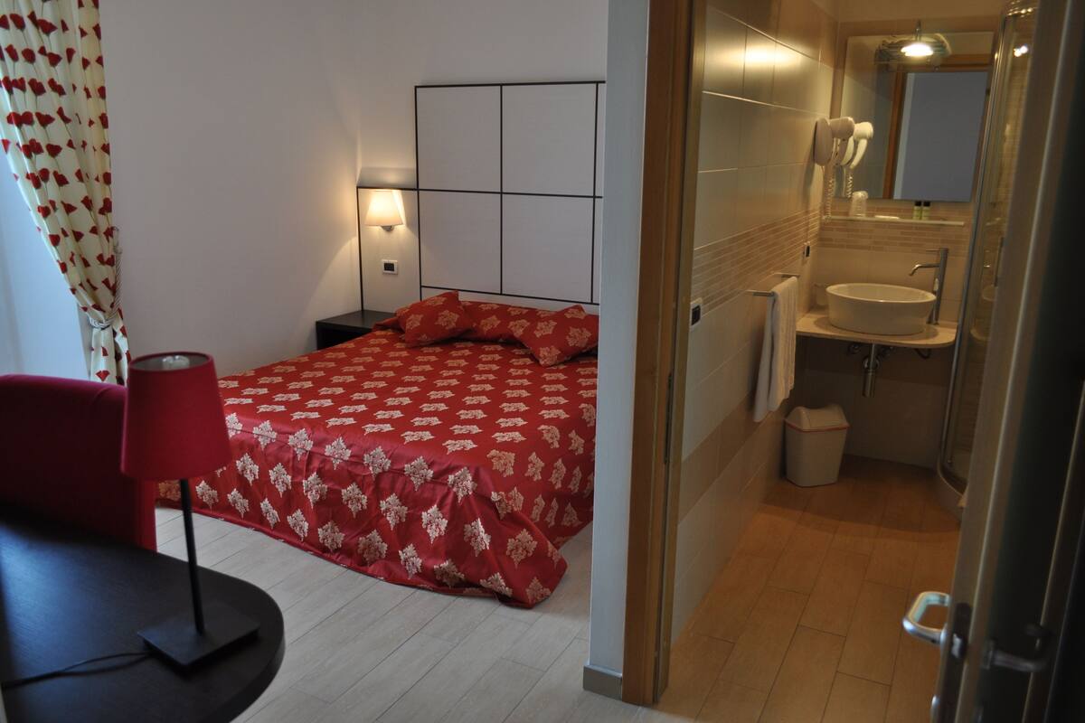 A hotel room with a bed, a bedside table, a lamp, a red chair, a bathroom with a sink and mirror, and a shower.