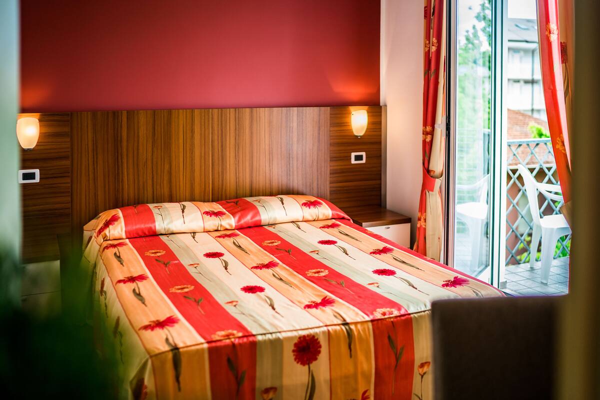 A hotel room with a wooden headboard, a bed with a floral bedspread, two wall-mounted lights, and a view of a balcony with a white chair.