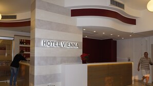 Una zona de recepción de hotel con diseño moderno, que incluye un mostrador de recepción, un letrero que dice "HOTEL VIENNA" y un jarrón decorativo con flores.