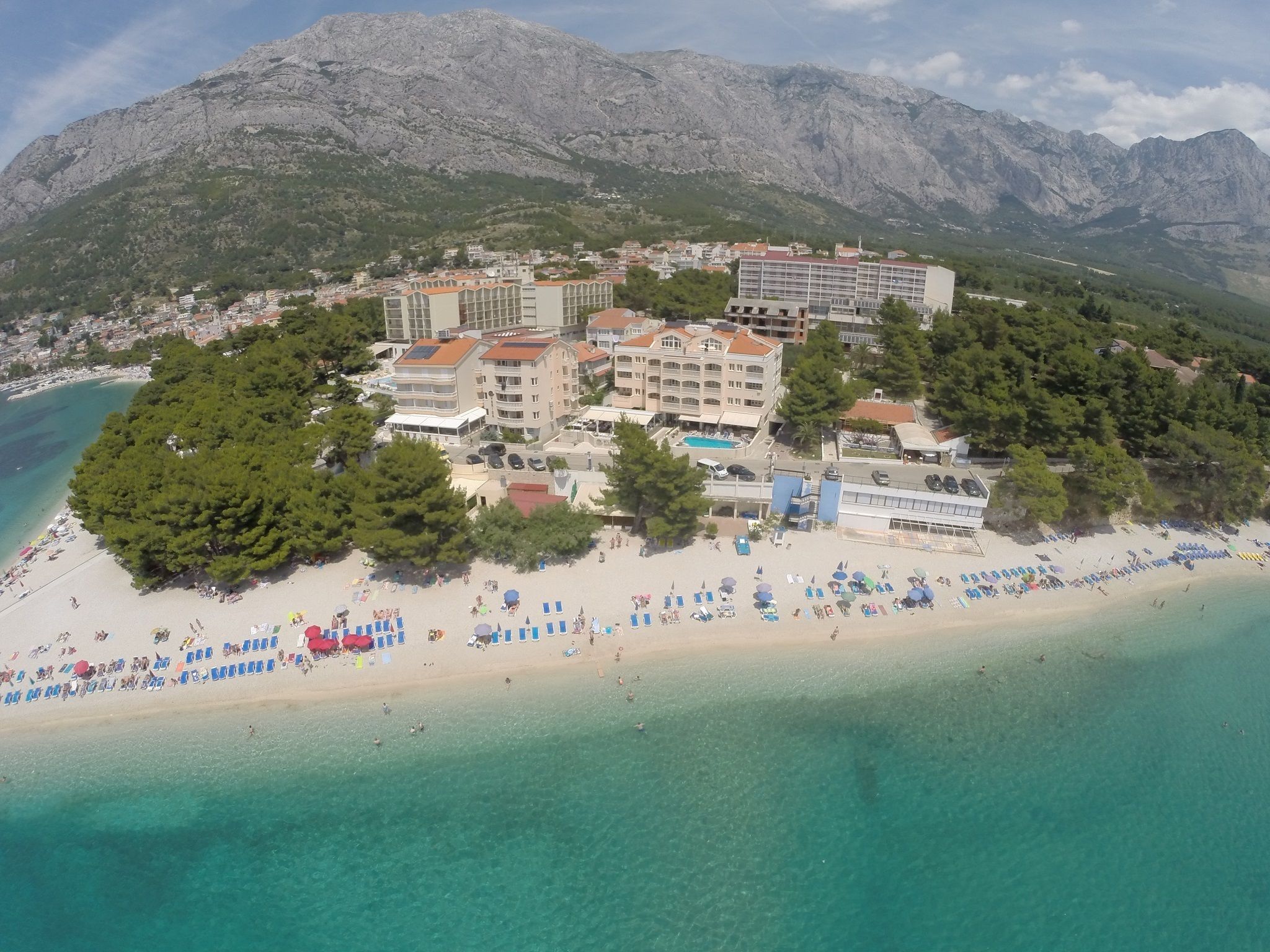 Vue aérienne d’un hôtel situé sur la côte, composé de plusieurs bâtiments, avec une plage et un paysage montagneux en arrière-plan.