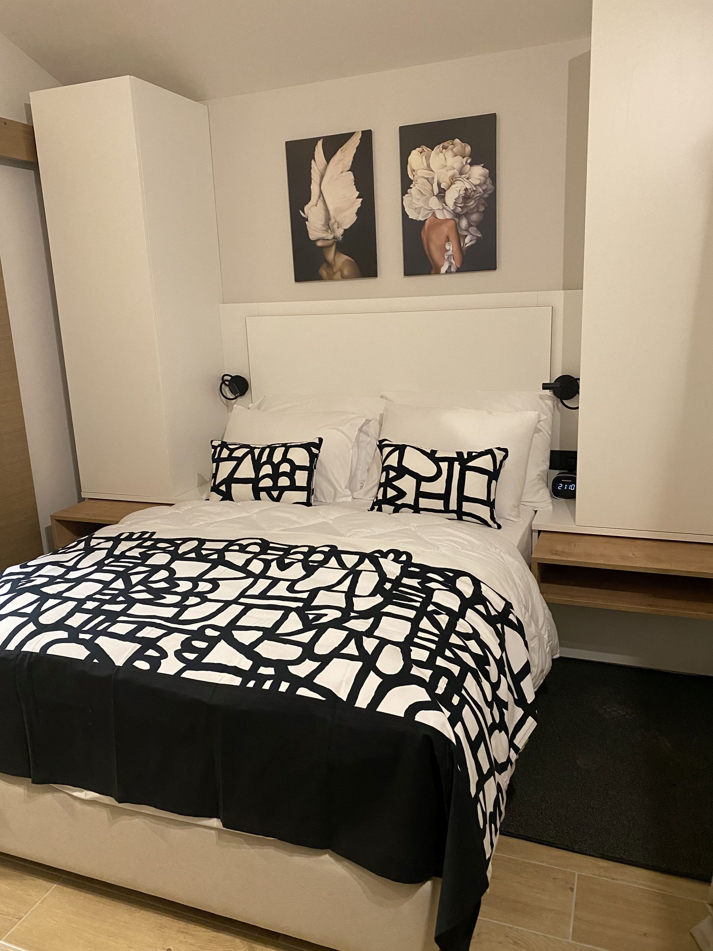 Un dormitorio con cama, cabecera blanca, dos relojes de pared y dos obras de arte enmarcadas sobre la cama.