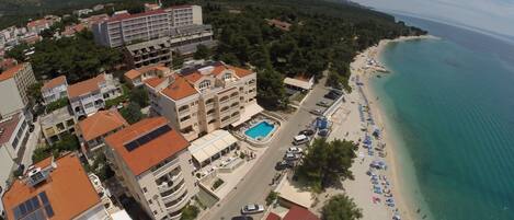 Vista aérea de una zona costera con playa, piscina y una cadena montañosa al fondo.