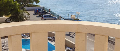 Un balcón con vista a una piscina, autos estacionados y una masa de agua distante.