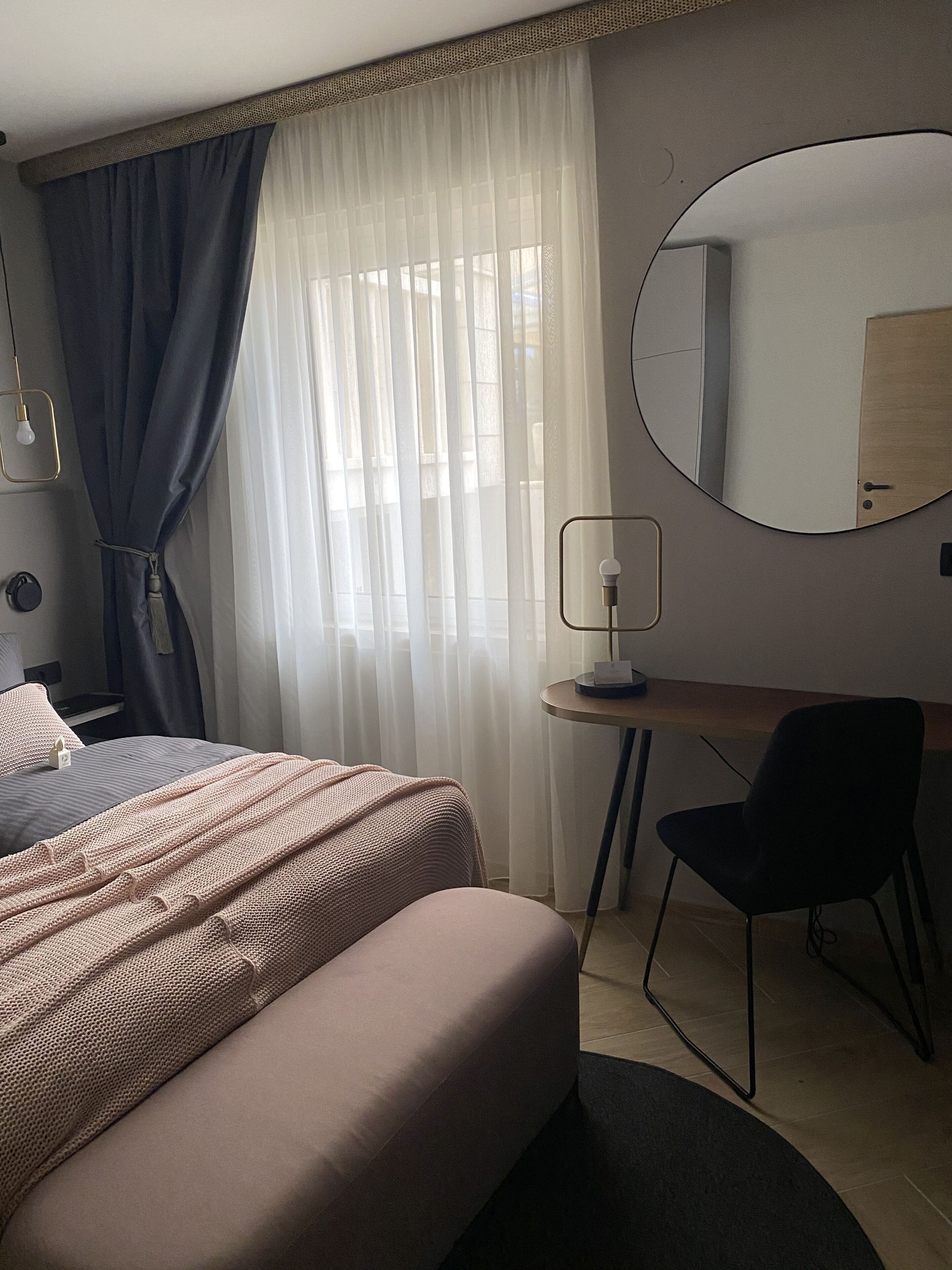Ein modernes Hotelzimmer mit einem Bett, einem Schreibtisch, einem Sessel und einem runden Spiegel, ausgestattet mit transparenten Vorhängen.