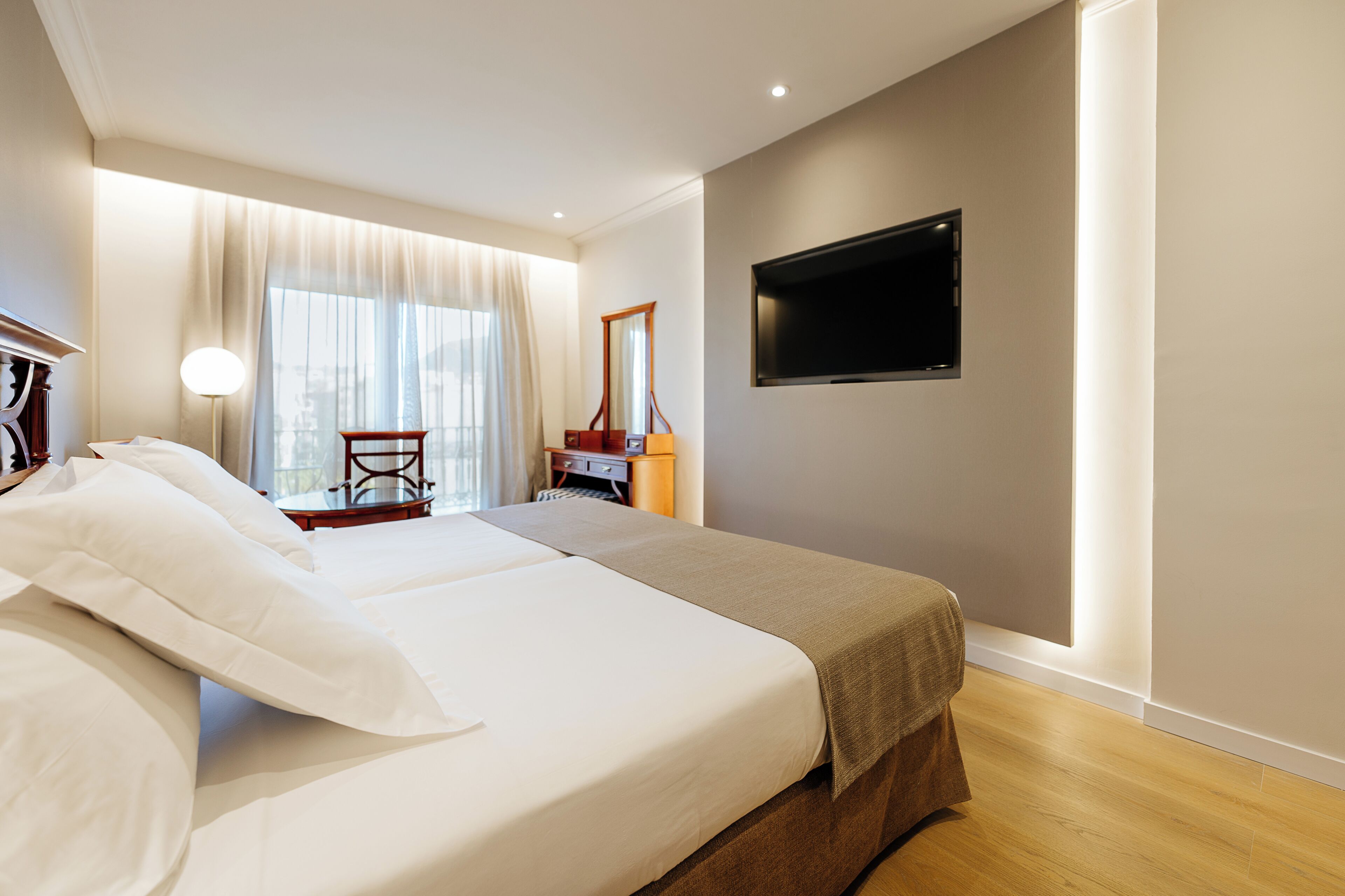 A hotel room with two beds, a desk, a chair, a television, and a large window with curtains.
