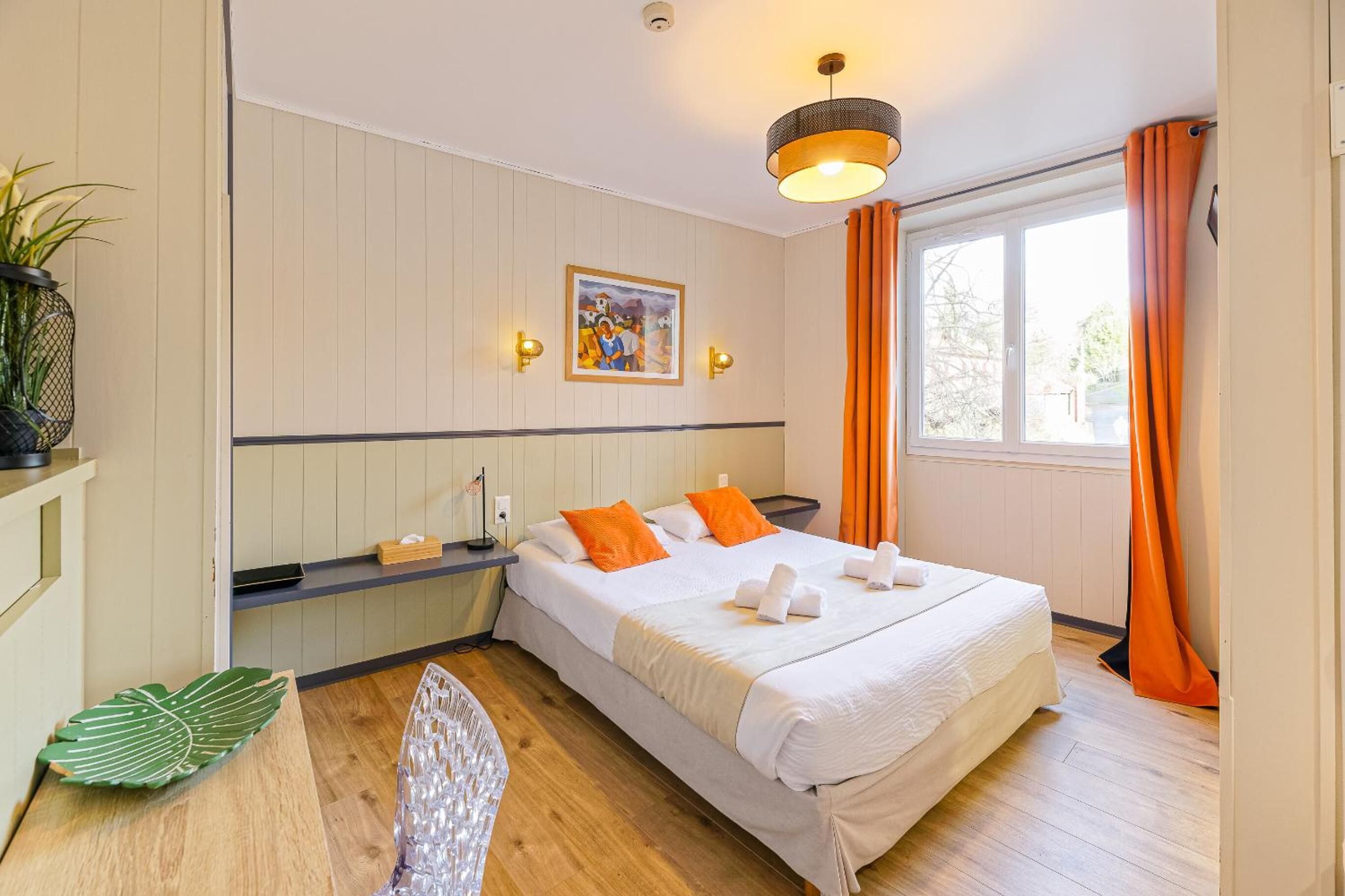 Une chambre d’hôtel avec un lit, un tableau, une fenêtre avec des rideaux, une plante et un bureau avec une chaise.
