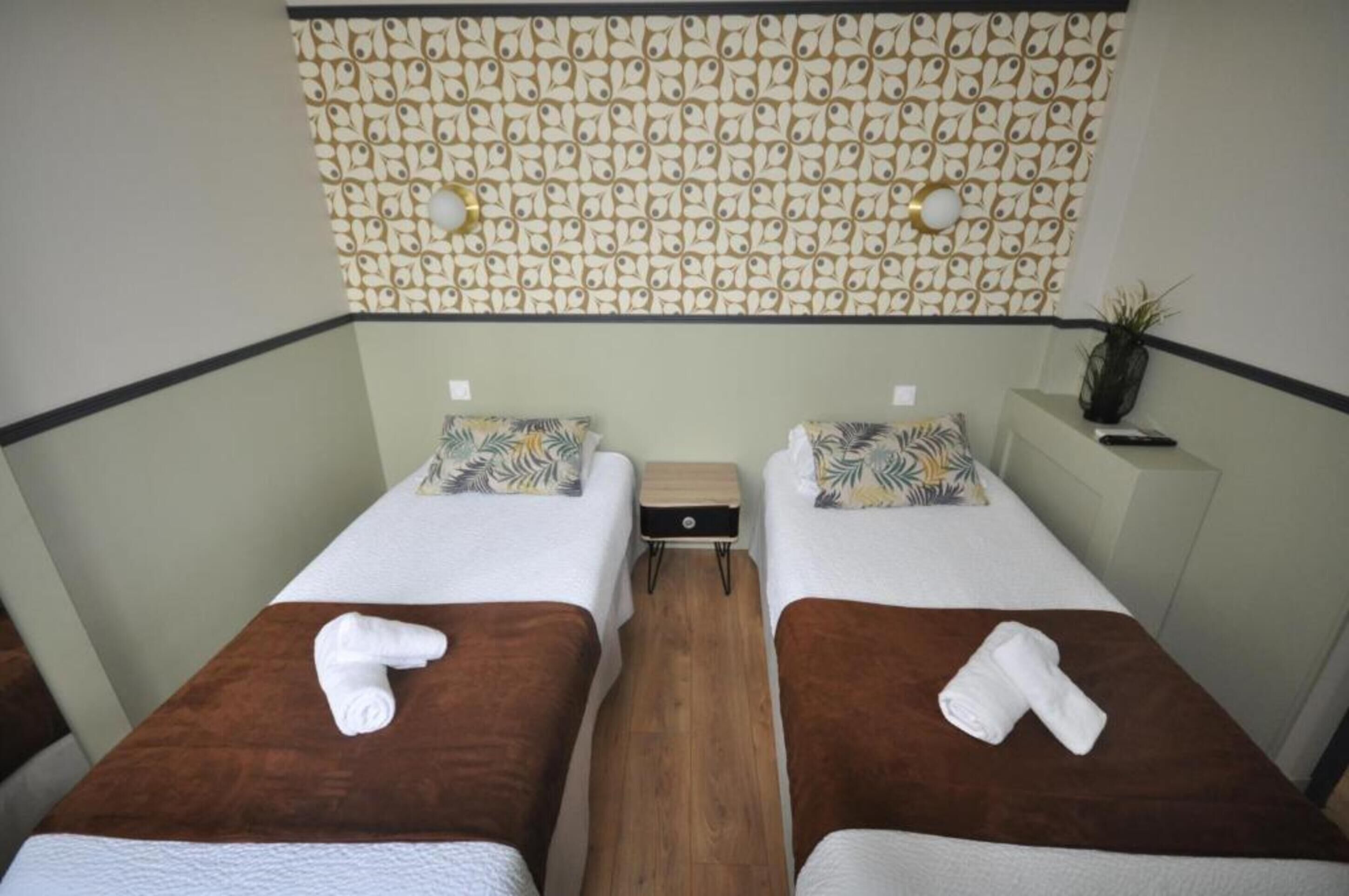 Una habitación con dos camas, cada una con cabecero marrón, ropa de cama blanca y una toalla sobre la cama. Hay una mesita de noche de madera entre las camas, y una pared con diseño de hojas sobre ellas.